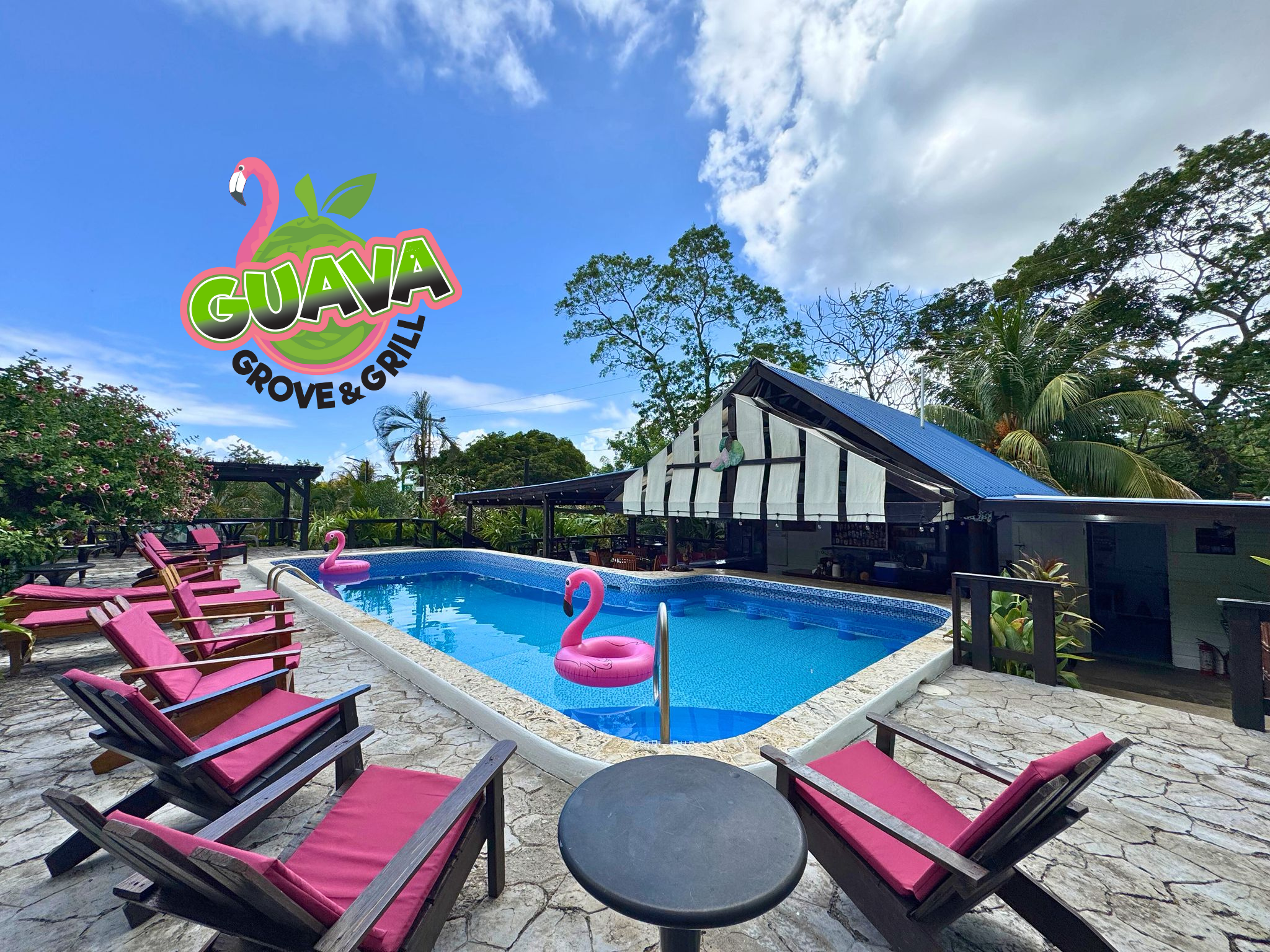 A tropical outdoor area featuring a swimming pool with pink flamingo floaties, surrounded by wooden lounge chairs with red cushions. In the background, a restaurant with a blue roof and lush greenery under a clear blue sky.